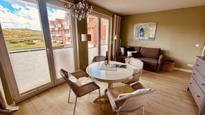 A room with a round table, wicker chairs, a sofa, a painting, and a view of buildings through large windows. - Im Dünengrund "Rebinka" DUE/01 - Apartment 02 - Dünengrund (Sylt)