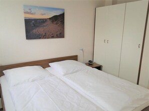 A hotel room with a bed, a nightstand with a lamp, a wardrobe, and a painting of a beach scene on the wall. - In the Dünengrund "Dünenrose" - Dünengrund (Sylt)