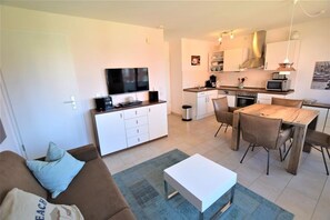A modern living room with a sofa, coffee table, and TV. Adjacent is a compact kitchen with a dining area. - In the Dünengrund "Dünenrose" - Dünengrund (Sylt)