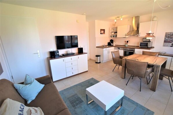 A modern living room with a sofa, coffee table, and TV. Adjacent is a compact kitchen with a dining area. - In the Dünengrund "Dünenrose" - Dünengrund (Sylt)