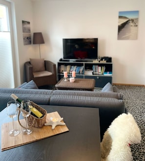 A living room with a sofa, a coffee table with wine glasses and a bottle, a television, and a bookshelf.