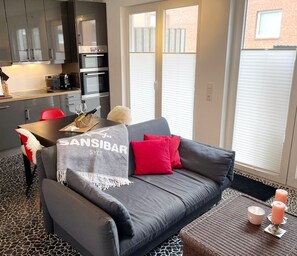 Een moderne woonkamer met een grijze bank, rode kussens, een tafel met een fles en glazen, en een keuken op de achtergrond.