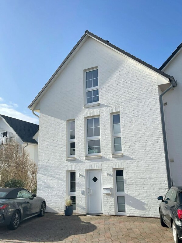 A white brick building with a central door, two windows on the ground floor, and a garage door. - House Dünenblick DUE/12 - Dünengrund (Sylt)