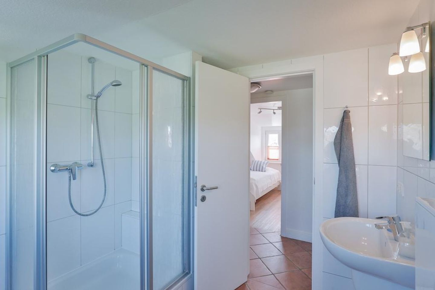 Un baño moderno con cabina de ducha de vidrio, un lavabo blanco y una habitación visible al fondo.