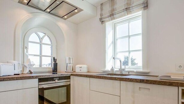 A modern kitchen with white cabinetry, a built-in oven, a large window, and a view of the outdoors. - Dünen Hüs II HO/34b - Dünen Hüs II (Sylt)