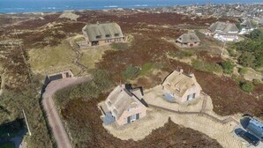 An aerial view of a coastal village with houses surrounded by natural terrain, a clear sky, and a view of the sea. - Dünen Hüs II HO/34b - Dünen Hüs II (Sylt)