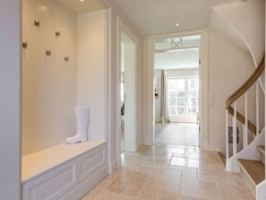 A spacious hallway with a white bench, a wooden staircase, and a view into a bright room with large windows.