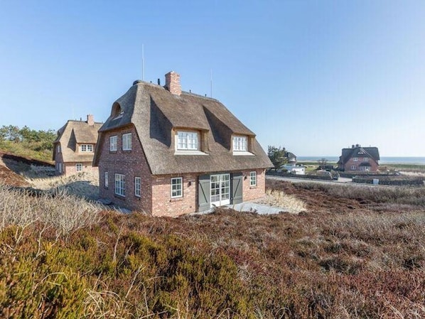 Une maison traditionnelle en briques, avec un toit de chaume, située sur une colline verdoyante et offrant une vue sur la mer.