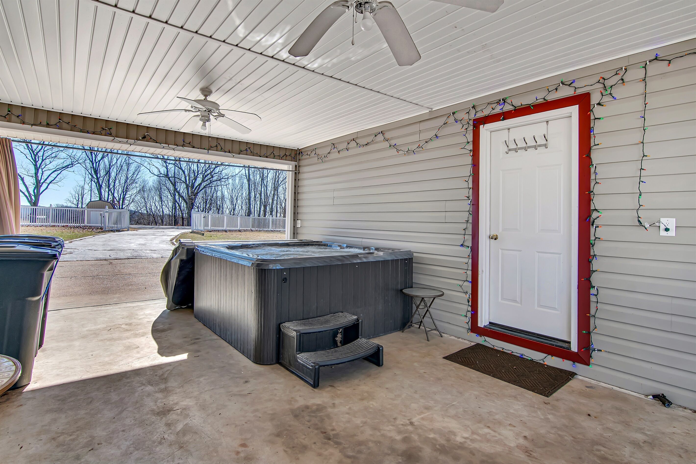 A covered outdoor area with a hot tub, a ceiling fan, and a door decorated with Christmas lights.