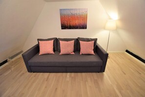 Ein dunkelgraues Sofa mit drei rosafarbenen Kissen in einem Raum mit Holzboden und einer Wandleuchte.