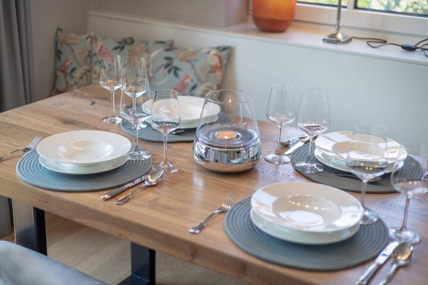 A dining table set with plates, glasses, and cutlery, with a window and a floral cushion in the background.