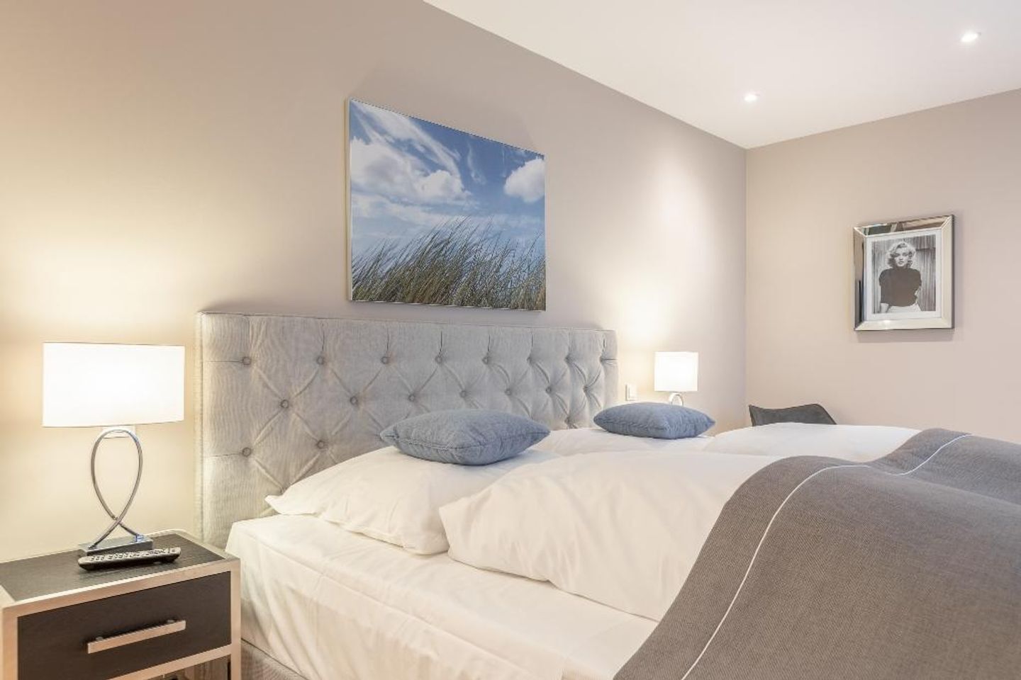 A bedroom with a tufted headboard, a large bed with white and gray bedding, two bedside lamps, a nightstand with a remote control, and a framed picture on the wall.