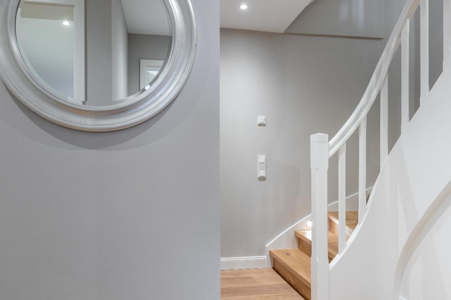 A staircase with wooden steps and white railings, leading to a landing with a light switch and a wall-mounted mirror.