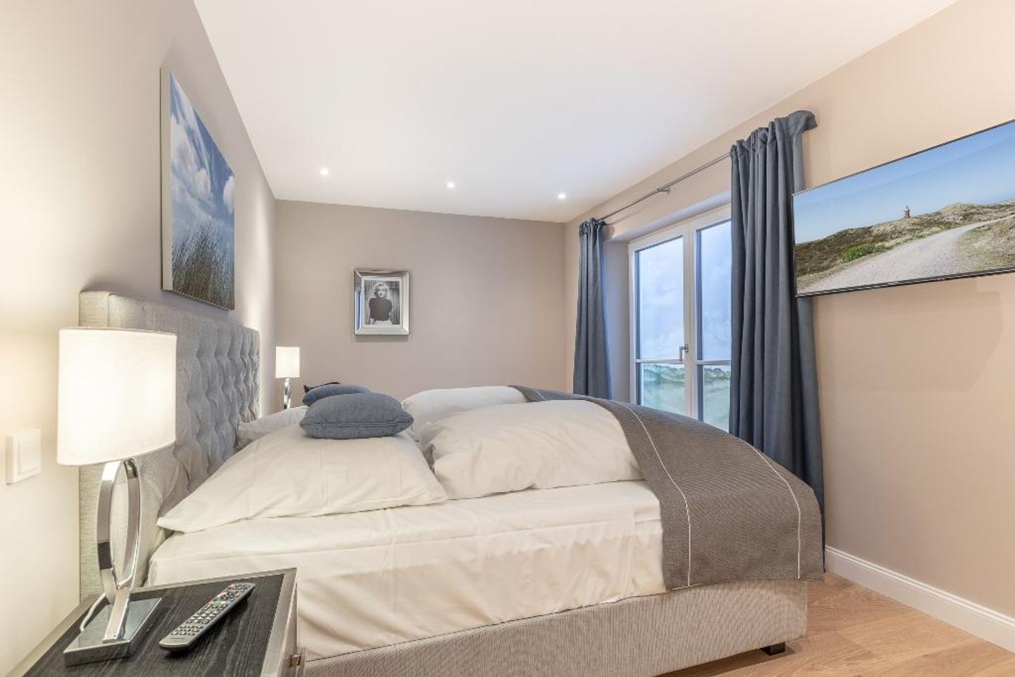 A modern hotel room with a curved bed, a bedside table with a lamp, a remote control, and a framed picture on the wall.
