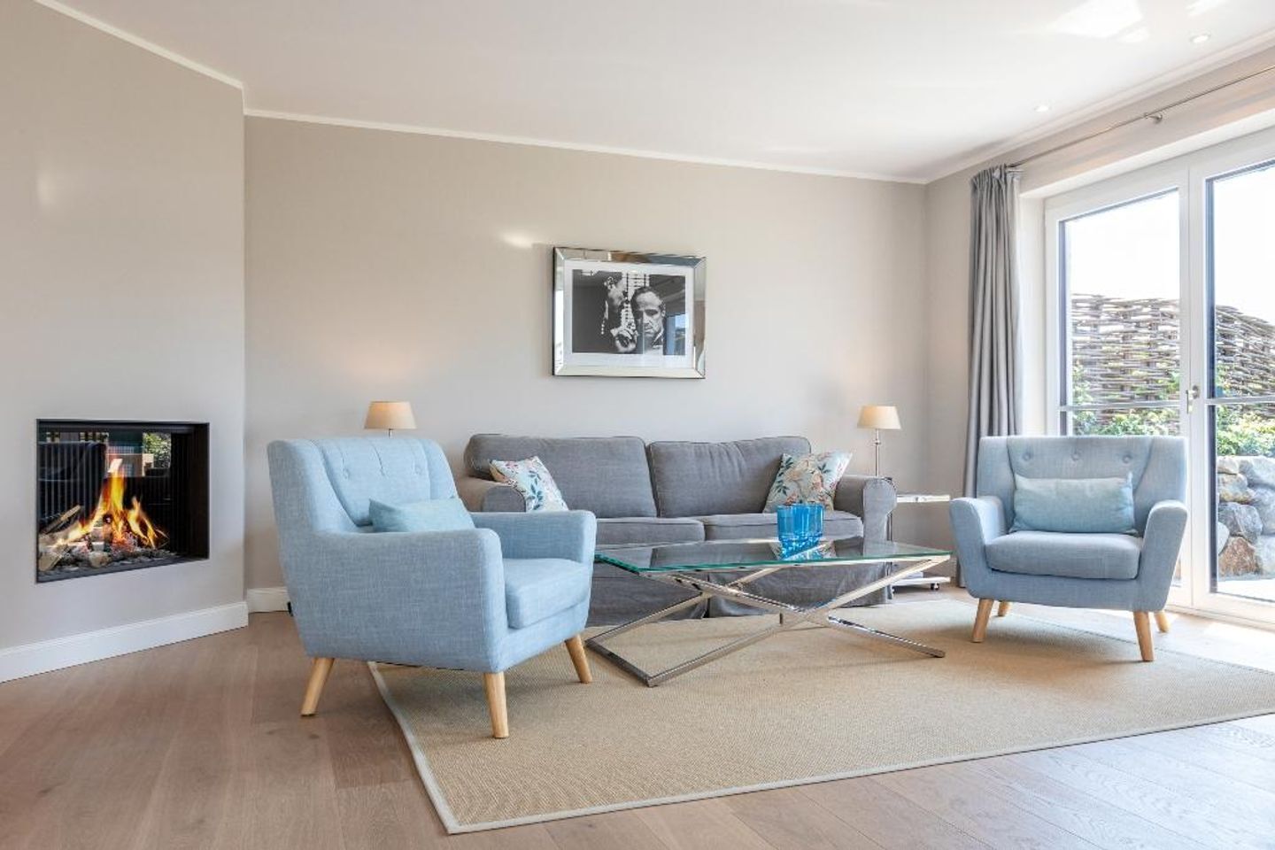 A modern living room with a fireplace, grey sofas, blue armchairs, and a glass coffee table.