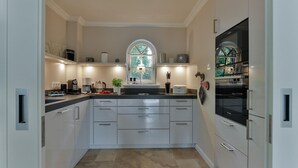 A modern kitchen with white cabinets, a central island, and a large window with a view of greenery. - "Raanwaikoje" RA/14B - Raanwaikoje (Sylt)