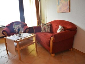 Una sala de estar con un sofá rojo, una mesa de centro de madera y una silla. Hay una ventana con cortinas y un cuadro en la pared.