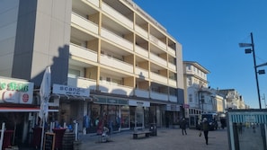 Ein mehrstöckiges Gebäude mit Balkonen, Geschäften darunter, darunter eine Apotheke und eine Galerie, sowie ein Fußgängerbereich mit einer Bushaltestelle.