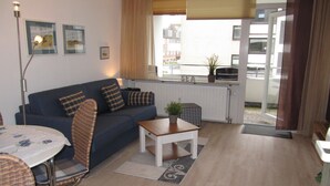 Un salon comprenant un canapé, une petite table, un fauteuil et une fenêtre donnant sur des immeubles.