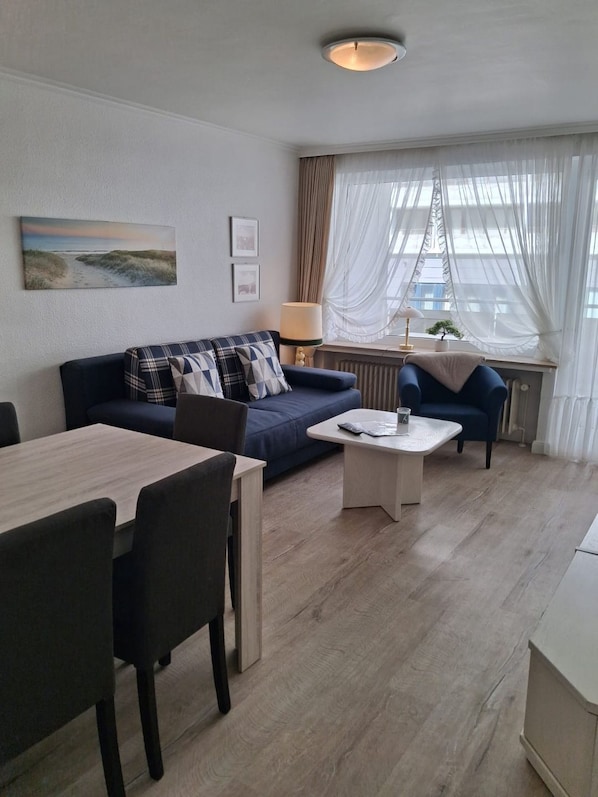 A modern living room with a sofa, a dining table, and a coffee table. There is a window with sheer curtains and a view of the outside.