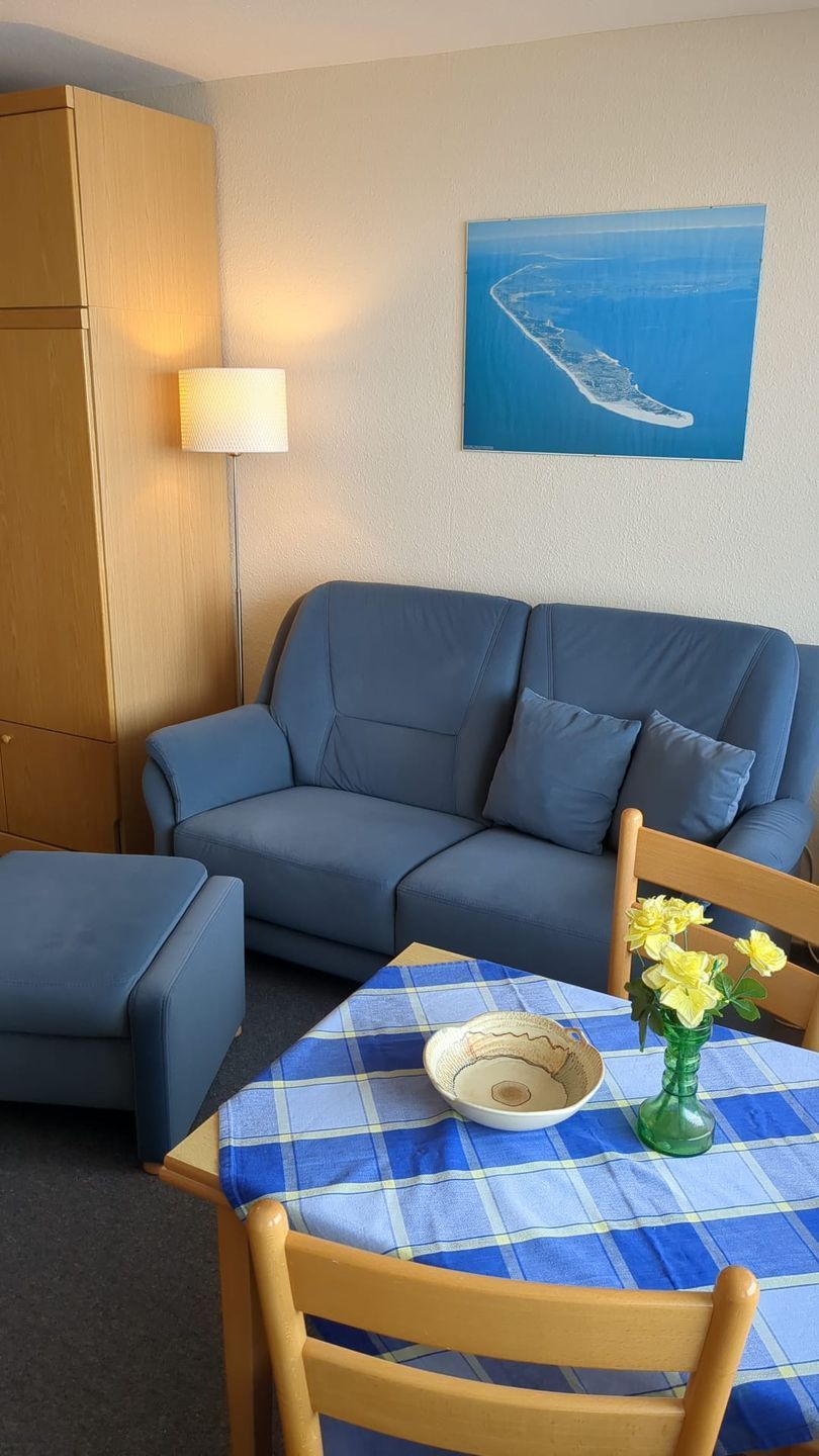 Une pièce avec un canapé bleu, un pouf assorti, une chaise en bois, une table recouverte d’une nappe à carreaux bleus et blancs, un vase avec des fleurs jaunes et une lampe fixée au mur.