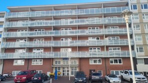 Un edificio residencial de varios pisos con balcones, una entrada visible y coches estacionados al frente.