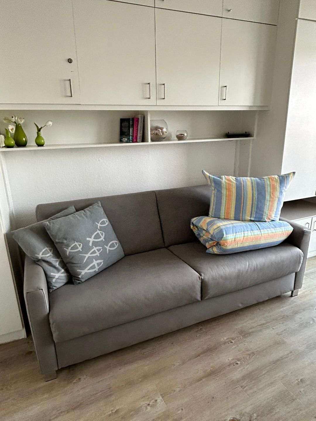 Un canapé en cuir gris, agrémenté de deux coussins : l’un avec un motif blanc et l’autre avec un motif rayé multicolore, dans une pièce dont le sol est recouvert de parquet et où se trouvent des meubles blancs en hauteur.