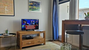 Un salon avec un meuble TV en bois, une table en verre ornée d’une plante, et une vue sur un immeuble à travers une fenêtre avec des rideaux bleus.