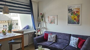Un salon moderne avec un canapé gris, une table à manger et une fenêtre avec des rideaux. Deux œuvres d’art encadrées ornent le mur.