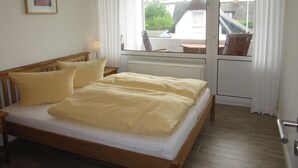 A bedroom with a wooden bed, a bedside table, a lamp, a window with curtains, and a view of a building outside.