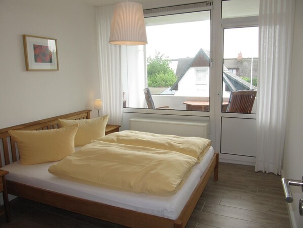 A bedroom with a wooden bed, a bedside table, a lamp, a window with curtains, and a view of a building outside. - Apartment Deckerstrasse 19-21 App. 17 - 3 room apartment in Deckerstr. (Sylt)