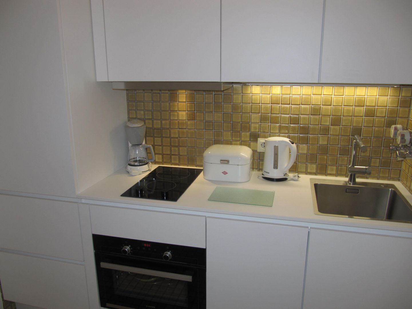 A compact kitchen with a built-in oven, a coffee maker, a kettle, and a sink.