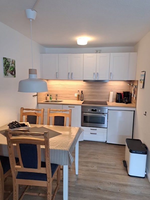 Une cuisine de taille réduite avec des meubles blancs, un plan de travail en bois, une table à manger avec des chaises et une poubelle.