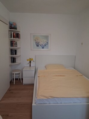 Ein Schlafzimmer mit einem Bett, einem Nachttisch, einem kleinen Tisch, einem Bücherregal und einem gerahmten Bild an der Wand.