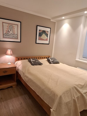 A bedroom with a wooden bed, two pillows, a nightstand with a lamp, and two framed artworks on the wall. - Halemdüür 9 An Apartment No. 1 - 3 room apartment in the south of Westerland. (Sylt)