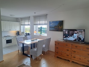 A modern kitchen with white cabinets, a dining table with chairs, a wooden cabinet with drawers, and a television displaying a cooking show. - Halemdüür 9 An Apartment No. 1 - 3 room apartment in the south of Westerland. (Sylt)