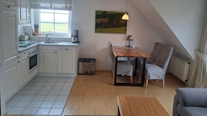 A compact kitchen with white cabinets, a sink, and a coffee maker. There's a dining table with chairs and a painting on the wall. - Halemdüür 9 An apartment no. 2 - 2 room apartment in the south of Westerland. (Sylt)