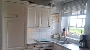 A kitchen with white cabinets, a stove, an oven, a sink, and a window with blinds. - Halemdüür 9 An apartment no. 2 - 2 room apartment in the south of Westerland. (Sylt)