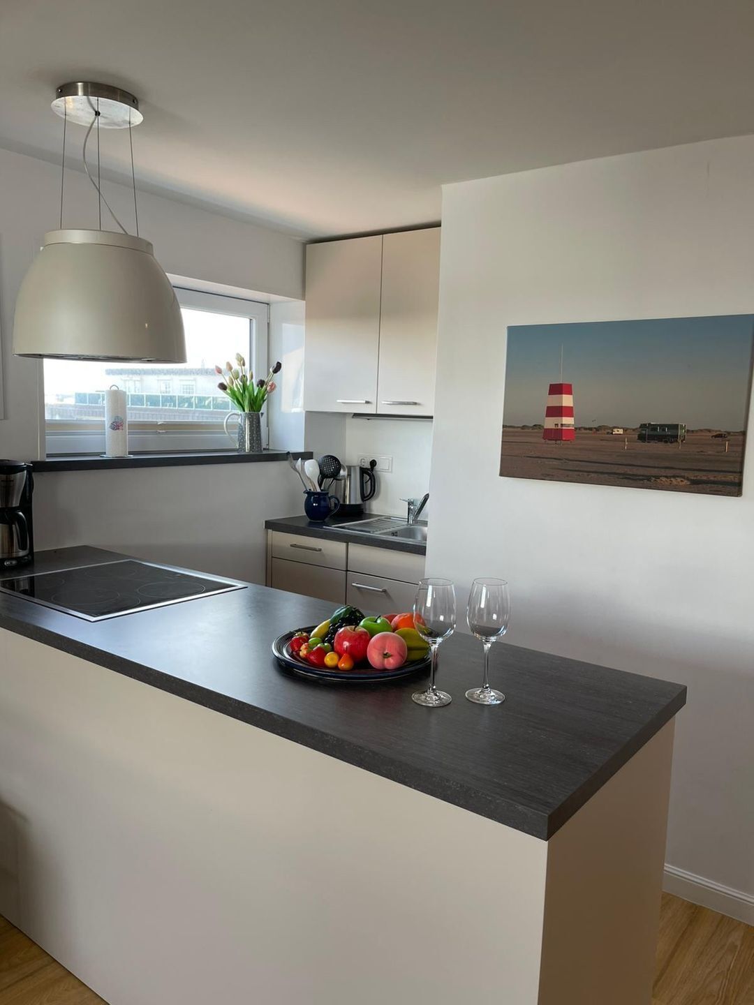 Une cuisine moderne avec un îlot central, une grande fenêtre et un tableau représentant un phare accroché au mur.