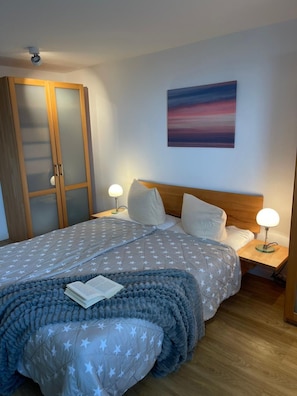 Un letto rifatto con cura, coperta a motivi stellati, due abat-jour, una testiera in legno, un quadro appeso alla parete e un armadio con ante in vetro satinato.
