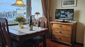 Een hotelkamer met een eettafel, gedekt met wijnglazen en een fles wijn, een televisietoestel dat een scène van de politie toont, en een houten dressoir.