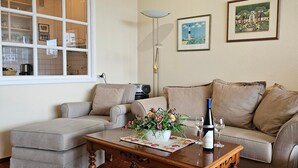 Een woonkamer met een bank, een voetenbankje en een salontafel. Er staat een lamp, er hangen afbeeldingen in lijsten aan de muur en op de tafel staat een fles wijn.
