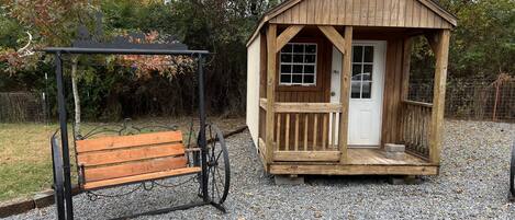 A wooden gazebo with a bench and a door, surrounded by gravel and trees.