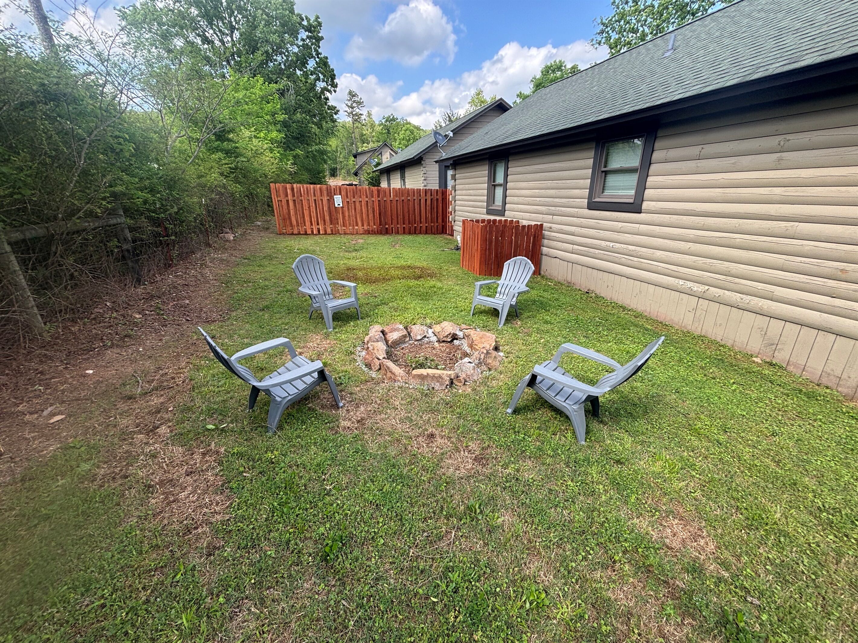 A backyard with a fire pit, four Adirondack chairs, a wooden fence, and a house with a satellite dish.