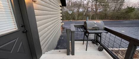 A wooden deck with a grill and a bench, a door with a doormat, and a railing overlooking a body of water.