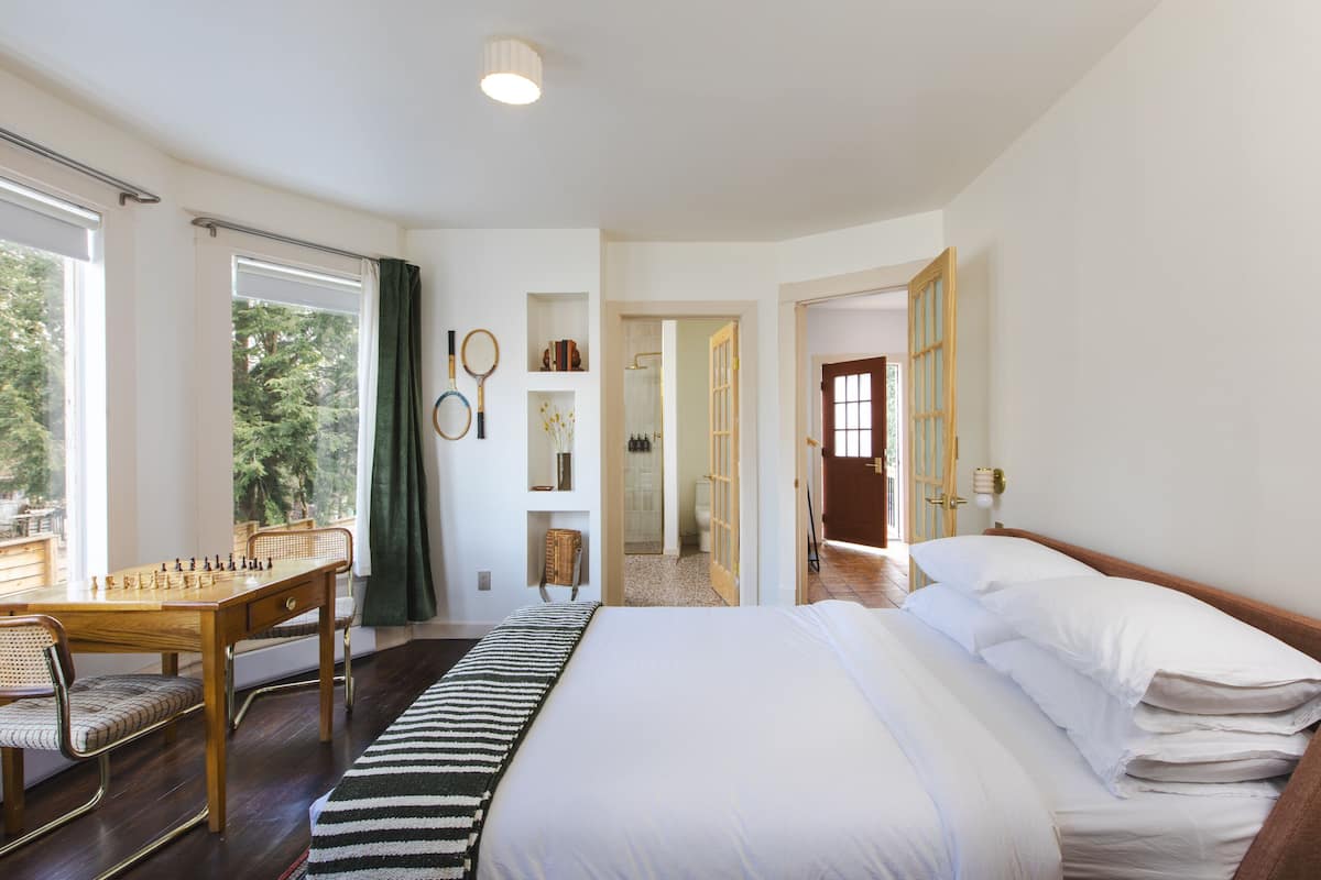 A bedroom with a large bed, a wooden desk with a chessboard, and a view of the outdoors through the windows.