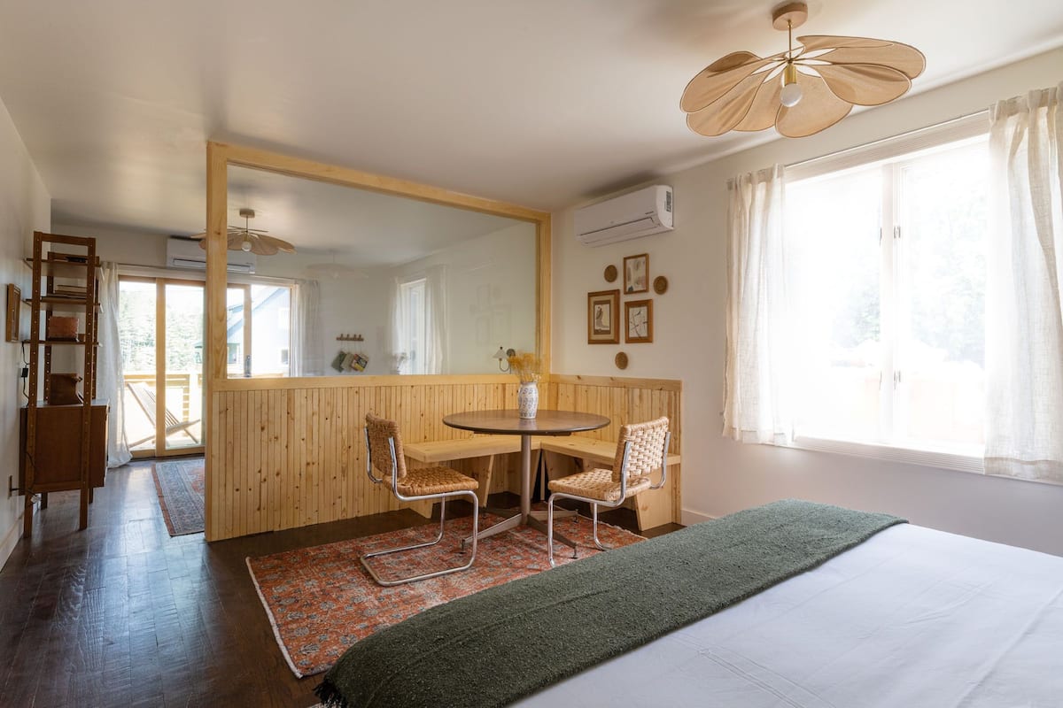 A room with a bed, a dining table with chairs, a large mirror, and a window with curtains.