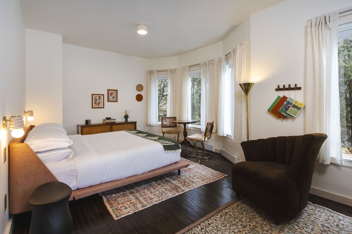 A bedroom with a bed, bedside table, chair, and a desk with a chair. There are windows with curtains, a wall-mounted shelf with books, and a floor lamp.