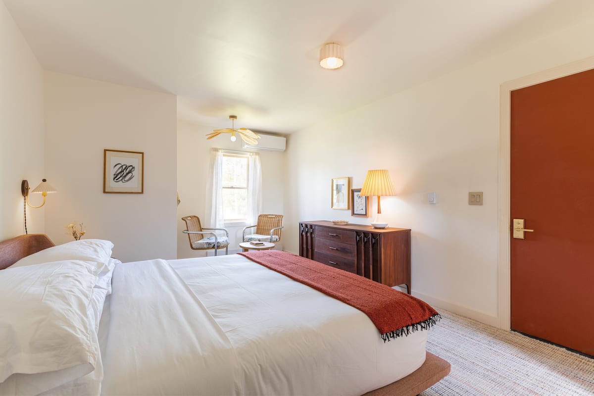 A bedroom with a large bed, a desk, a chair, a lamp, and a door.
