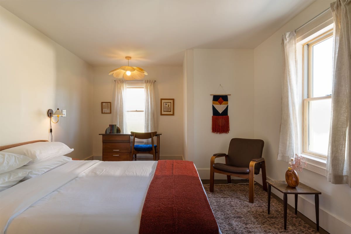 A hotel room with a bed, a desk, a chair, a small table with a vase, and a window with curtains.